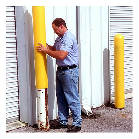 A man in a light blue shirt and jeans secures an UltraTech Post Protector 152_ from UltraTech International, a vibrant yellow polyethylene sleeve, over a metal post near the white building with roll-up doors. In the background, another identical post is waiting for its protective covering.