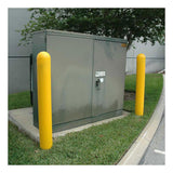 A green electrical utility box rests on a concrete slab, accompanied by two yellow UltraTech Post Protector 152 bollards from UltraTech International. Located near a gray building wall, the box is surrounded by grass at its base, with a portion of the road visible to the right.