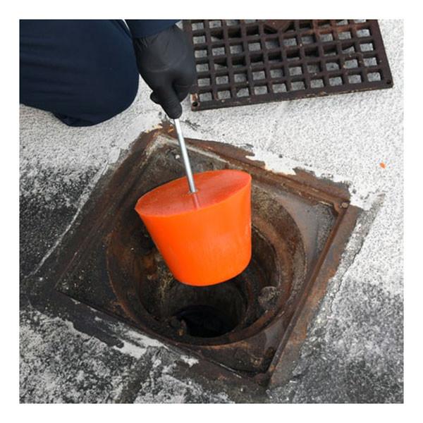 A person wearing black gloves is placing an UltraTech Drain Plug 211 made by UltraTech International, crafted from flexible polyurethane material, into a square metal storm drain on a cement surface. The plug, connected to a metal rod, acts as an efficient spill response tool. Beside the drain opening lies a square grate.