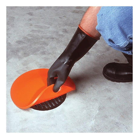 A person wearing black rubber gloves is placing an UltraTech Drain Seal 213_ from UltraTech International over a floor drain. The flexible cover is specifically designed to block the drain. The person is kneeling on a gray concrete surface and is wearing blue jeans.