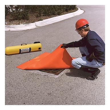 A worker wearing a red hard hat and gloves positions an UltraTech Drain Seal 213, from UltraTech International, over a storm drain on a grey asphalt road. A yellow cylindrical object is prominently visible nearby. In the background, shrubs and concrete borders form part of an effective stormwater management program.