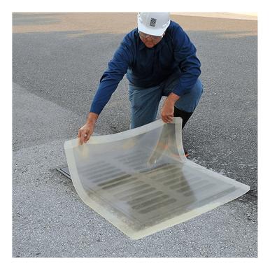 A person wearing a blue shirt and cap is kneeling on the pavement, adjusting an UltraTech Clear Model Drain Seal 220_ over a storm drain. This clear plastic cover from UltraTech International is designed for spill response and features a grid pattern to ensure it fits securely over the drain.