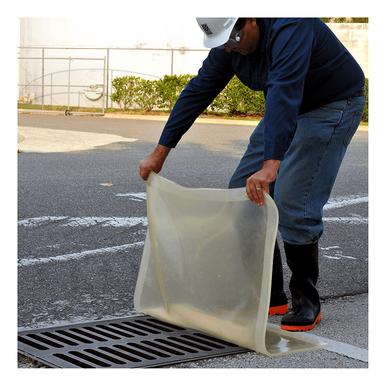 Wearing a construction hat and boots, an individual places an UltraTech Clear Model Drain Seal 220_ over a street drain. This spill response drain cover from UltraTech International enhances safety in the outdoor area, with a road and fence visible in the background.