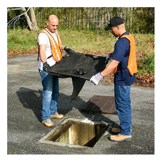 Two men wearing orange safety vests and gloves are carefully positioning an UltraTech International UltraTech Drain Guard Adjustable Frame Model 8930 over an open storm drain on a paved surface. In the background, lush grass and towering trees create a serene atmosphere around their focused task.