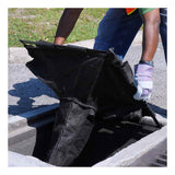 Dressed in a green shirt, jeans, and safety vest, an individual wearing gloves efficiently installs an UltraTech International UltraTech Drain Guard Adjustable Frame Model 8930 into a storm drain on a street. The large black filter, crafted from thick material, fits securely within its adjustable frame as grass waves gently in the background.