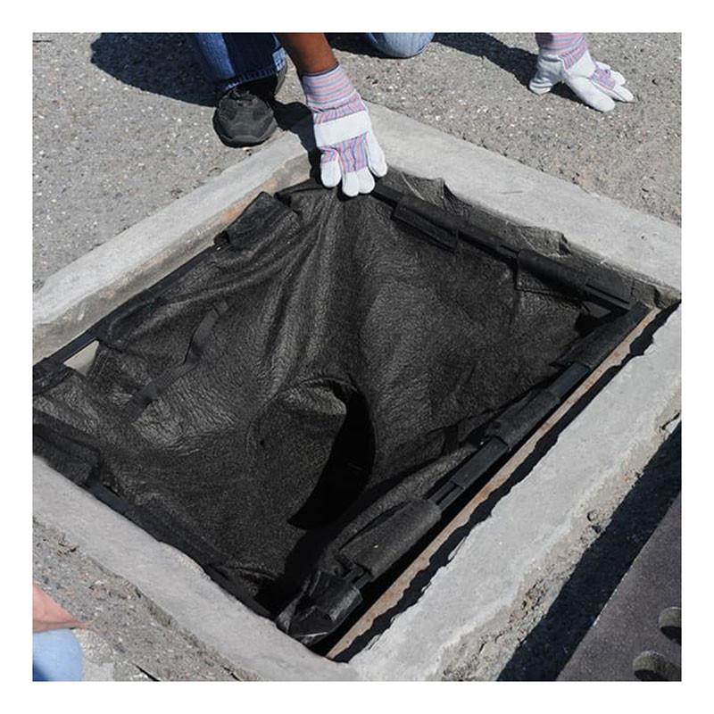 Two individuals are installing an UltraTech International Drain Guard Adjustable Frame Model 8930 with a black fabric catch basin insert into a square storm drain on a paved surface. One person is wearing white gloves with blue cuffs, and only their hands and lower legs are visible as they adjust the steel framework for a perfect fit.