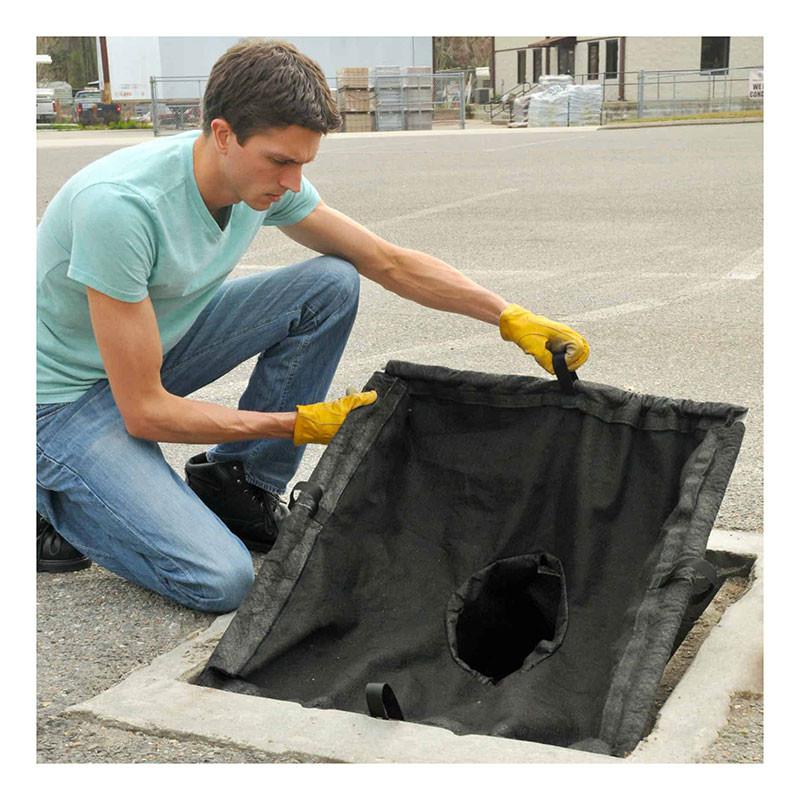 A man in a light blue shirt and yellow gloves lifts an UltraTech International UltraTech Drain Guard Adjustable Frame Model 8930 from a rectangular storm drain on a paved surface. Buildings and industrial materials are visible in the background.