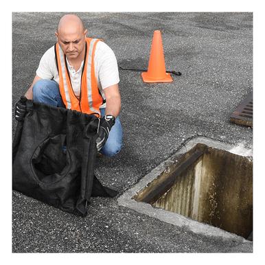 A person wearing an orange safety vest and gloves is kneeling beside an open storm drain on a paved surface, installing an UltraTech Economy Frame Model Drain Guard 8935- from UltraTech International. An orange traffic cone stands nearby.