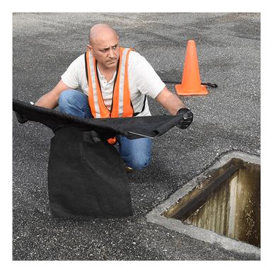 A man in an orange safety vest inspects a storm drain on a paved surface. He's holding an UltraTech Economy Frame Model Drain Guard 8935- near the opening, ensuring everything is secure. An orange traffic cone is placed beside the drain as he meticulously checks for any issues.