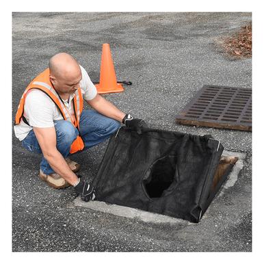 A worker wearing a safety vest is positioning an UltraTech Economy Frame Model Drain Guard 8935 over a storm drain on an asphalt surface, with an orange traffic cone located nearby.