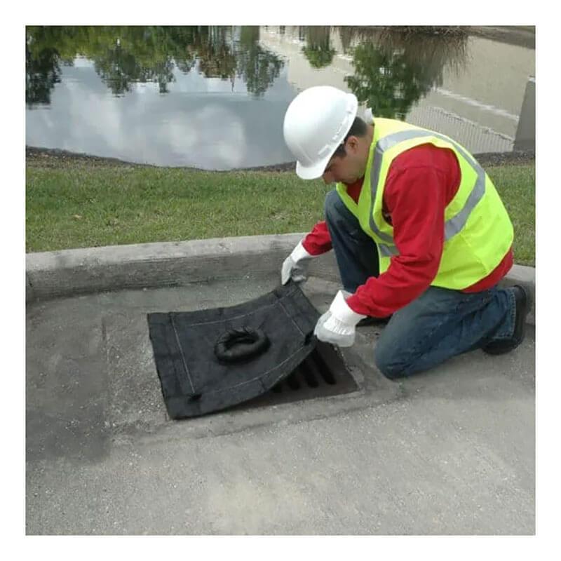 A worker wearing a white hard hat, yellow safety vest, red shirt, and jeans kneels on a concrete sidewalk to fit an UltraTech Inlet Guard Plus 916_ from UltraTech International over a storm drain. Nearby, the grassy area frames a small body of water that reflects trees and clouds.