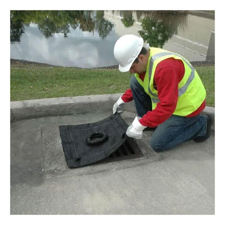 An employee in a white hard hat, yellow safety vest, and red shirt efficiently places an UltraTech Inlet Guard Plus 916_ over a storm drain using geotextile material on the concrete sidewalk. A pond with trees is reflected in the water nearby.