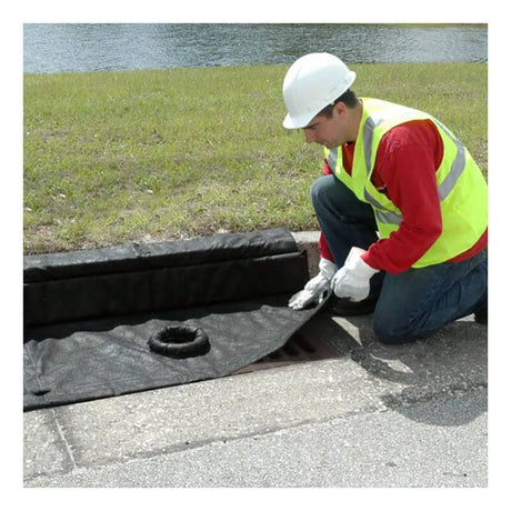 A worker wearing a white helmet and reflective vest kneels by a storm drain, positioning the UltraTech Inlet Guard Plus 916_ over it. This essential drain protection product from UltraTech International is located outdoors near a grassy area.