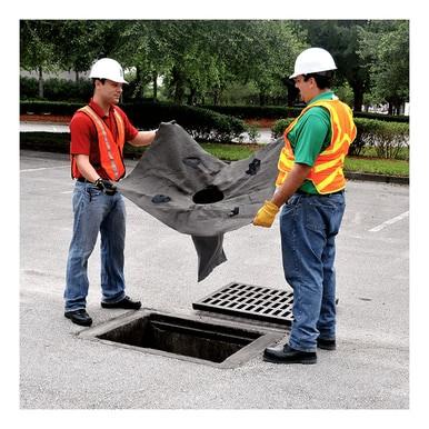 Two workers wearing hard hats and safety vests hold an UltraTech International product, the Oil and Sediment Drain Guard 921_ (10-Pack), over a storm drain in a parking lot, preparing to install the filter. The surrounding trees and greenery emphasize the importance of managing stormwater runoff.