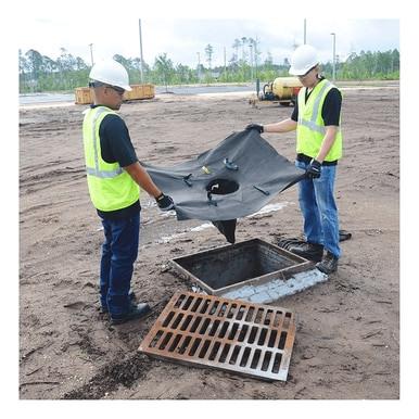 Two construction workers in safety gear position an UltraTech International Oil and Sediment Drain Guard 921_ (10-Pack) over an open storm drain at a construction site, ensuring it filters stormwater runoff. A metal grate is placed nearby on the ground.