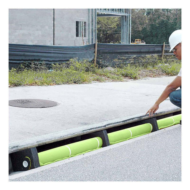 A worker wearing a hard hat is inspecting a storm drain on a construction site, equipped with the UltraTech Curb Guard Plus Model for stormwater runoff protection. Nearby are a sidewalk, grass, and a partially constructed building, all well-maintained thanks to the UltraTech International system in place.