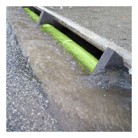 Rainwater flows along a street gutter, partially blocked by a bright green UltraTech Curb Guard Plus Model from UltraTech International, serving as a curb guard. The water is muddy, indicating heavy rainfall, and the pavement and road surface are visible.