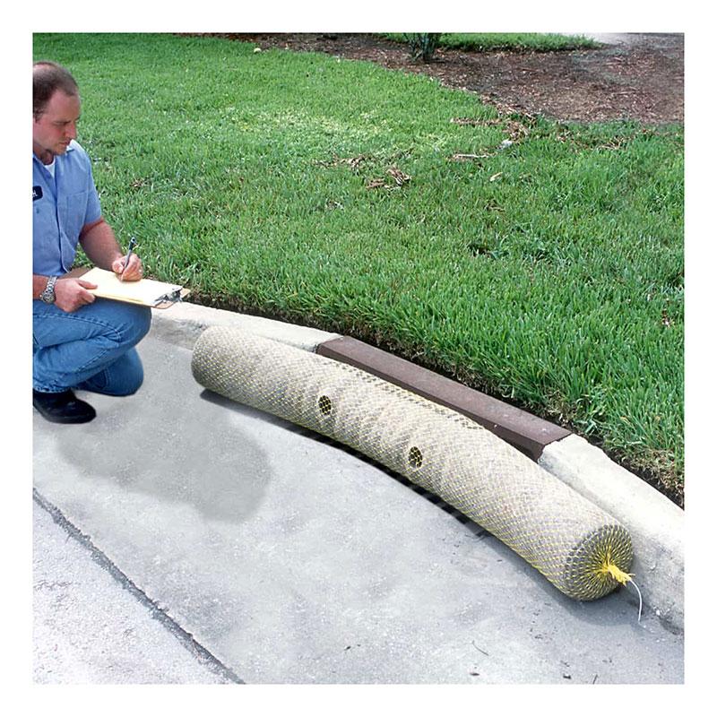 An individual kneels on a sidewalk, jotting notes on a clipboard. Next to them, adjacent to a grassy area, lies an UltraTech Gutter Guard by UltraTech International—a large, mesh-covered erosion control sock crafted from recycled synthetic fibers—demonstrating an innovative approach to stormwater protection.