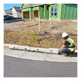 A construction worker in a hard hat and safety vest measures an UltraTech Gutter Guard on a curb in front of a partially constructed building with green sheathing. Focused on stormwater management, he ensures the UltraTech International curb inlet filters are correctly positioned amid the dirt and wooden debris scattered around the site.