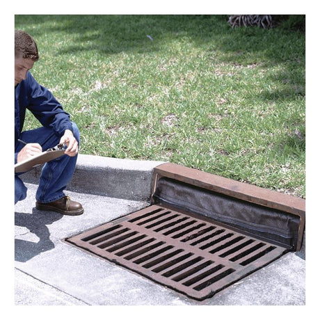 A person in a blue jacket crouches on a sidewalk, writing on a clipboard. Next to them, an UltraTech Reusable Model Drain Guard by UltraTech International is nestled among grassy vegetation, highlighting the focus on inspecting the catch basin insert for optimal functionality.