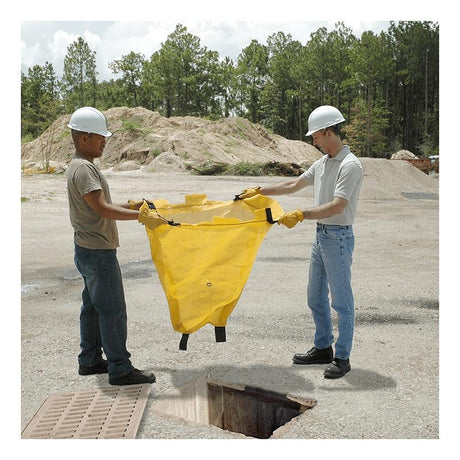 Two workers in hard hats and gloves carefully position an UltraTech Reusable Model Drain Guard by UltraTech International over an open manhole on the concrete surface. In the background, piles of sand and a line of trees stretch into the distance.