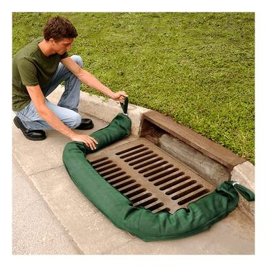 A person kneels beside a storm drain on a street curb, adjusting an UltraTech Filter Sock 945_ from UltraTech International, placed in front of the drain. This setup, essential for stormwater protection, is surrounded by grass and pavement.