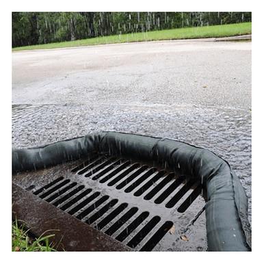 Rainwater flows into a street storm drain, encircled by a protective barrier equipped with UltraTech International's UltraTech Filter Socks 945_ for enhanced pollutant removal. Wet pavement and a grassy area are visible in the background, emphasizing the dedication to stormwater protection.