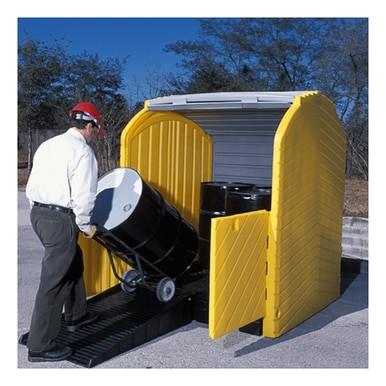 A person wearing a red helmet and safety goggles carefully maneuvers a large black barrel on a dolly into an outdoor storage unit with open doors. The UltraTech Hard Top P4 Plus Spill Pallet 963, in its yellow design and set on a paved area, offers reliable drum containment against the scenic backdrop of trees.