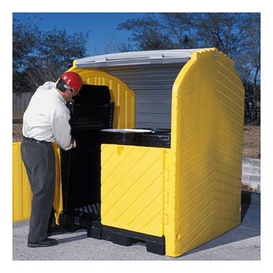 A person wearing a red hard hat and protective eyewear opens an UltraTech Hard Top P4 Plus Spill Pallet 963 by UltraTech International, a large yellow outdoor storage container designed for drum containment. The container is situated on a concrete surface, with lush trees visible in the background.