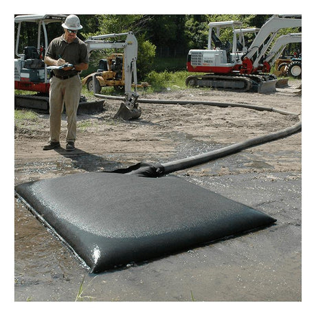 A worker in a hard hat checks a tablet at the construction site, where an UltraTech Dewatering Bag 972_-OS from UltraTech International is placed on muddy ground. It's connected to a hose for effective sediment filtration, with excavators visible in the background amidst greenery to ensure storm water pollution prevention is maintained.