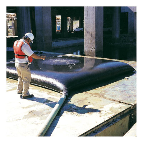 A person wearing a white hard hat, safety goggles, and an orange safety vest inspects the large black UltraTech Dewatering Bag 972-OS by UltraTech International on a construction site platform. A hose is attached to the bag for sediment filtration, with concrete structures visible in the background.