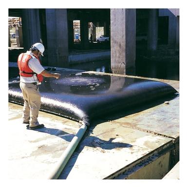 A construction worker, wearing a white hard hat and an orange safety vest, inspects a large black UltraTech Dewatering Bag 972_-OS from UltraTech International at the site. This essential tool for sediment filtration is connected to a hose, with concrete columns rising in the background as evidence of successful storm water pollution prevention measures.