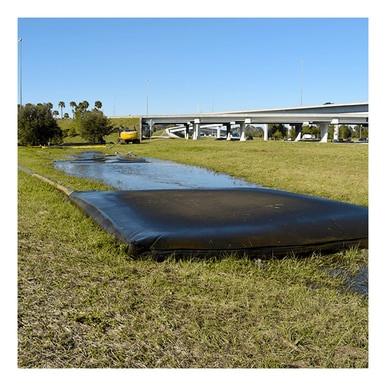 An UltraTech Dewatering Bag 972_-OS from UltraTech International rests on a grassy field, bordered by shallow water, functioning as part of a storm water pollution prevention system. In the background, there's a highway overpass and trees under a clear blue sky.