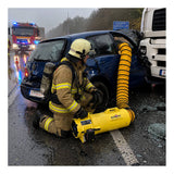 A firefighter kneels beside a crashed car and truck in the rain, operating a yellow Euramco RAMFAN UB20 18VDC with Quick-Couple Canister for jobsite ventilation, as emergency vehicles with flashing lights are visible in the background.