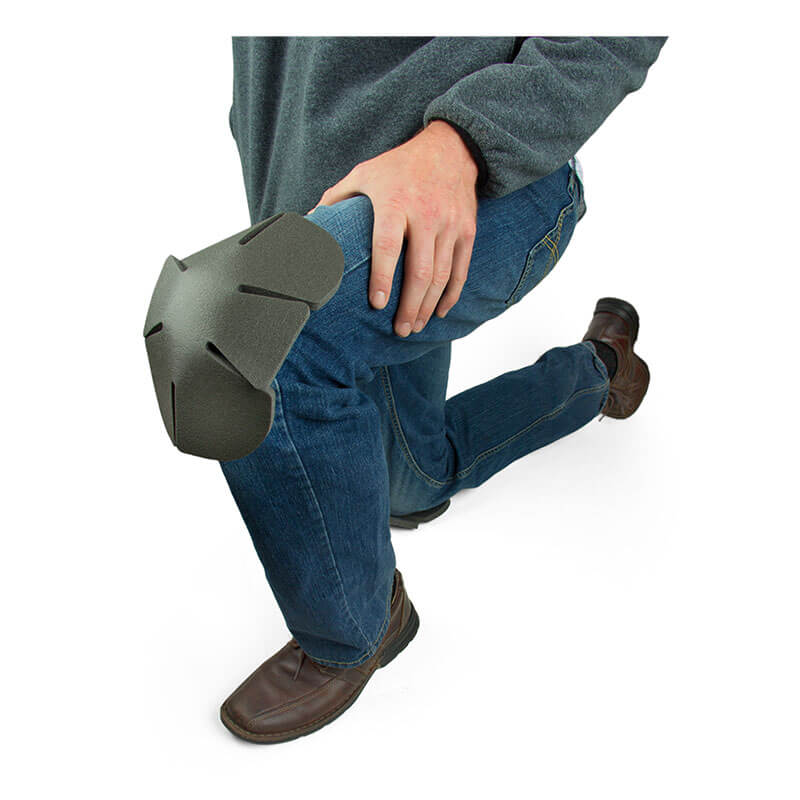 A person kneels on the ground, wearing blue jeans and brown shoes. They have a gray Working Concepts Soft Knees Disposable Knee Pad 1012 on their knee, with their hands resting on this protective garment. They are dressed in a dark sweatshirt.