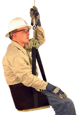 A person, equipped with a hard hat, safety glasses, and gloves, sits confidently in a French Creek Deluxe Work Seat 4153 by French Creek Production. Wearing a beige shirt and jeans, they seem prepared for work at height or in a safety training environment, highlighting strong fall protection measures.