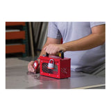 A person in a light blue shirt is handling an Accuform STOPOUT Pry-Resistant Lock Box KCC690 by AccuformNMC on a table. The lock box is red and has multiple locks attached to it, while the individual is holding a padlock. Shelves with various objects can be seen in the background.