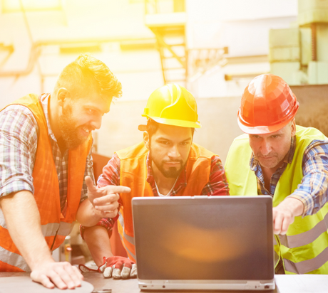 Three construction workers wearing safety vests and helmets are gathered around a laptop, focusing intently on the screen. They appear to be discussing or analyzing something. The background shows industrial equipment and a warm lighting effect.