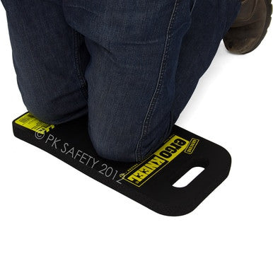 A person is kneeling on the ergonomic foam rubber mat labeled "Working Concepts ErgoKneel Kneeling Mat 5040 8 x 16 in." by Working Concepts. The black mat, with a handle cutout on one end, provides support for someone wearing dark blue jeans and brown work boots.