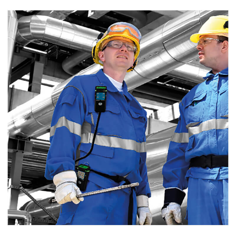 Two workers in blue protective uniforms, gloves, and helmets stand in an industrial facility with metal pipes in the background. Both wear safety goggles and communication devices clipped to their uniforms, each equipped with a GfG G999S 4 Gas Detector with Integrated Pump from GfG to monitor for multi-gas hazards during confined space entry.