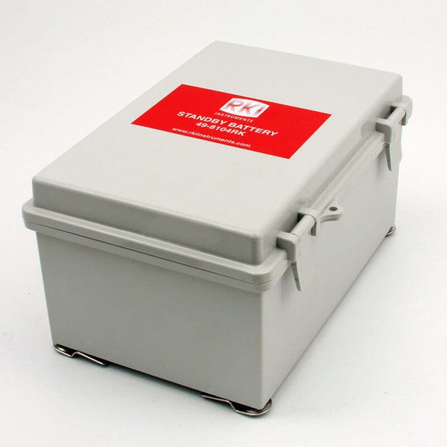 A rectangular gray box featuring a red label on the lid displays "RKI Instruments Standby Battery Assembly, 24 VDC, 12 AMP Hours 49-8104RK." This battery assembly is equipped with two clasps to secure the lid and sits firmly on a metal base, making it ideal for powering beacon applications.