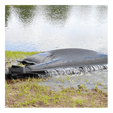 An UltraTech Dewatering Bag 972_-OS by UltraTech sits near a pond’s edge, releasing filtered water onto grass—an effective solution for stormwater pollution prevention. Trees reflect in the pond behind it.