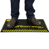 A person wearing muddy boots stands on the Working Concepts ErgoKneel Extreme Standing Mat 5020, measuring 16 x 28 inches—perfect for workers who need to stand for extended periods. This anti-fatigue mat offers a textured surface along with caution stripes around the edges, ensuring comfort and safety in challenging settings.