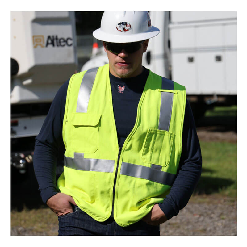 A person wears an NSA DRIFIRE FR Lightweight Long Sleeve T-Shirt DF2-CM-446LS, a white hard hat, sunglasses, and a yellow reflective safety vest while standing outdoors near utility vehicles with hands in pockets.