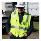 A person wears an NSA DRIFIRE FR Lightweight Long Sleeve T-Shirt DF2-CM-446LS, a white hard hat, sunglasses, and a yellow reflective safety vest while standing outdoors near utility vehicles with hands in pockets.