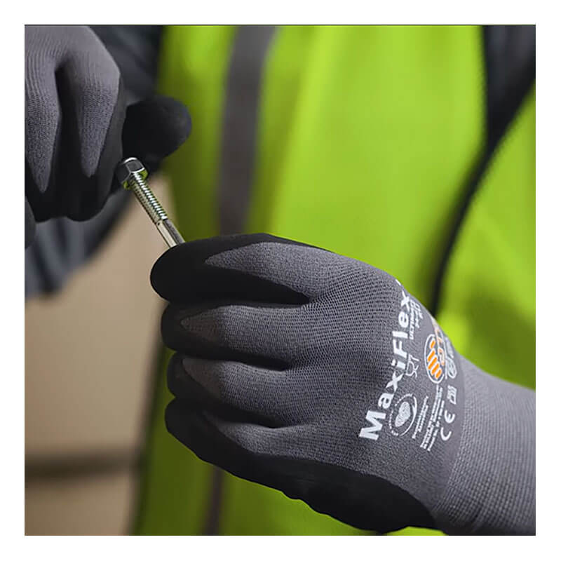 A person in a yellow safety vest is holding a screw or bolt with both hands while wearing PIP ATG MaxiFlex Ultimate Micro-Foam 3/4 Coated Palm Nitrile Gloves (34-875).