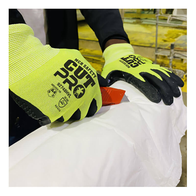 A worker wearing MCR Safety Cut Pro 18 Ga HyperMax Hi Vis Nitrile Micro Dotted Gloves (92748HV) uses a red utility knife to cut white material, possibly in an industrial or warehouse setting.