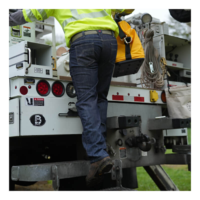 A worker in a high-visibility jacket and NSA DRIFIRE Relaxed Straight Leg FR Jean PNT-JRS02 climbs onto a utility truck while carrying a yellow tool bag.