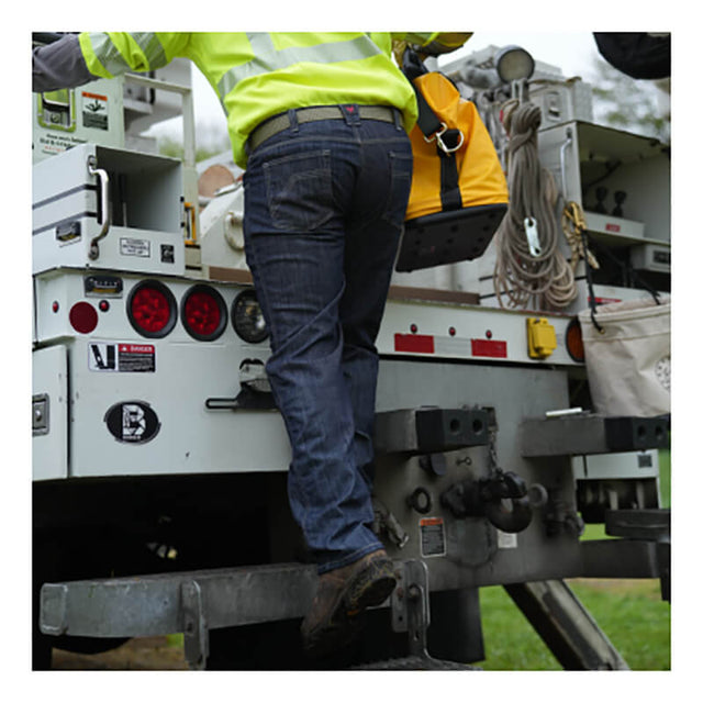 A worker in a high-visibility jacket and NSA DRIFIRE Relaxed Straight Leg FR Jean PNT-JRS02 climbs onto a utility truck while carrying a yellow tool bag.