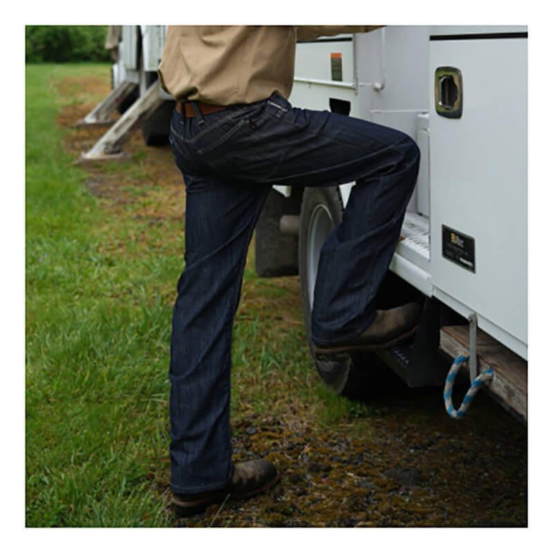 Wearing NSA’s DRIFIRE Slim Straight Leg FR Jean (PNT-JSS01) and sturdy work boots, a person steps onto a vehicle’s side step while holding the door. In the background, grass and other vehicles are visible.